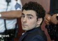 Luigi Mangione, dressed in a dark suit and blue chequered shirt, looks at the camera with law enforcement officials in the background
