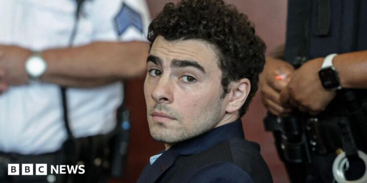 Luigi Mangione, dressed in a dark suit and blue chequered shirt, looks at the camera with law enforcement officials in the background