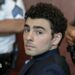 Luigi Mangione, dressed in a dark suit and blue chequered shirt, looks at the camera with law enforcement officials in the background