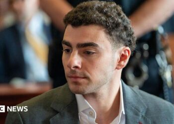 Luigi Mangione is seen from the shoulders up, sitting at the defence table in court. He wears a charcoal grey suit.