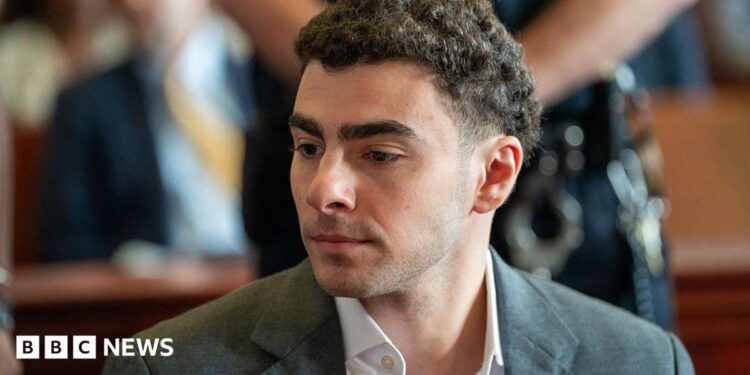 Luigi Mangione is seen from the shoulders up, sitting at the defence table in court. He wears a charcoal grey suit.