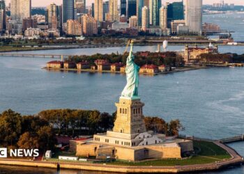 aerial shot of Statue of Liberty and waters surrounding it with New York skyline in background