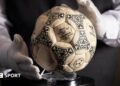 A person wearing white gloves places the football used in the 1986 World Cup quarter-final on a stand