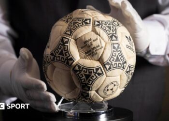 A person wearing white gloves places the football used in the 1986 World Cup quarter-final on a stand