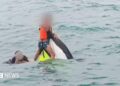 A boy wearing a lifejacket holds on to the top of a capsized jet ski as a man swims next to him in the sea
