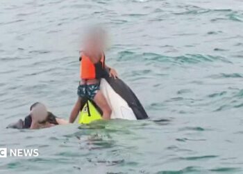 A boy wearing a lifejacket holds on to the top of a capsized jet ski as a man swims next to him in the sea