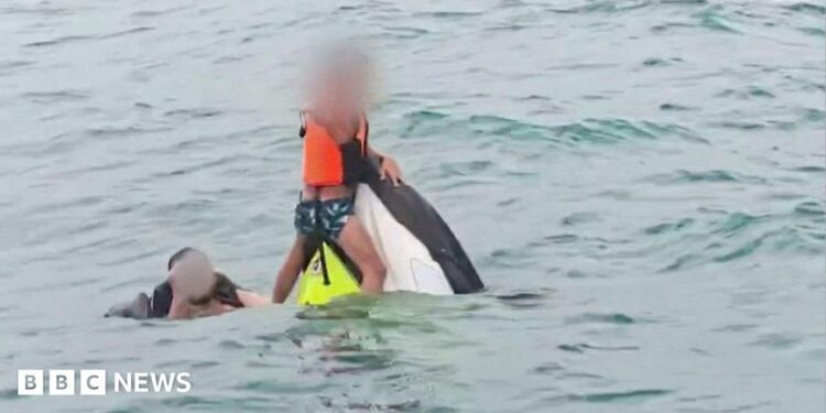 A boy wearing a lifejacket holds on to the top of a capsized jet ski as a man swims next to him in the sea