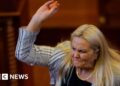 Blond woman wearing white and black blazer lifting right arm and holding egg
