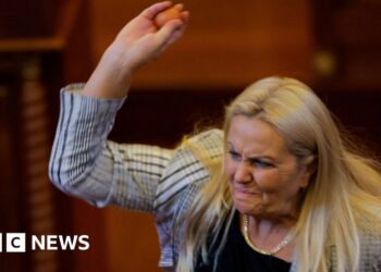Blond woman wearing white and black blazer lifting right arm and holding egg