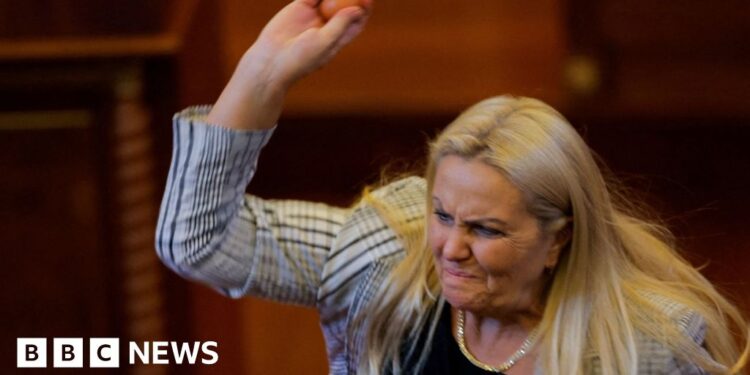 Blond woman wearing white and black blazer lifting right arm and holding egg