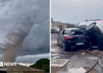 A tornado is seen spinning above a house in the clouds, with two cars on top of each other on the right.