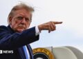 Donald Trump lifts his right arm up and points with his index finger. Part of the presidential seal can be seen behind him. He is looking to his right at the camera.