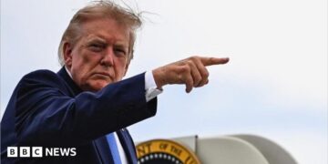 Donald Trump lifts his right arm up and points with his index finger. Part of the presidential seal can be seen behind him. He is looking to his right at the camera.
