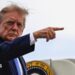 Donald Trump lifts his right arm up and points with his index finger. Part of the presidential seal can be seen behind him. He is looking to his right at the camera.