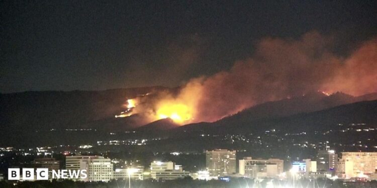 Two people look over what appears to be a domestic fence. They are looking at where smoke is rising from hills in the background.