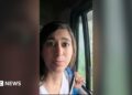A woman speaks to the camera as she sits in a moving car