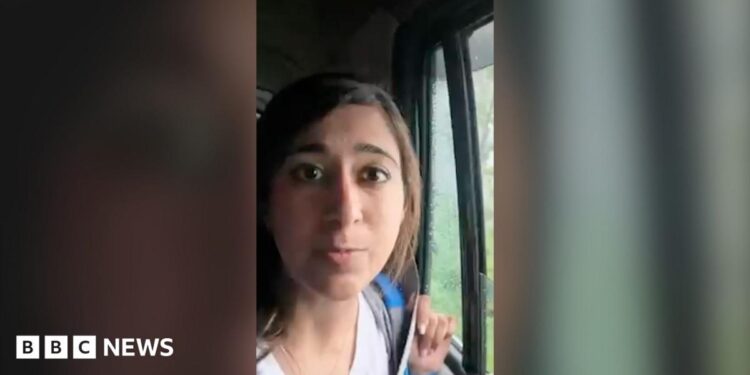 A woman speaks to the camera as she sits in a moving car