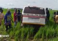 A group of men in Nigeria gather around a white vehicle that is stuck in a muddy rural area surrounded by long grass