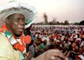An archive picture of Olusegun Obasanjo from 1999. He is wearing a large white hat and addressing a crowd of people through a red microphone that he is holding.