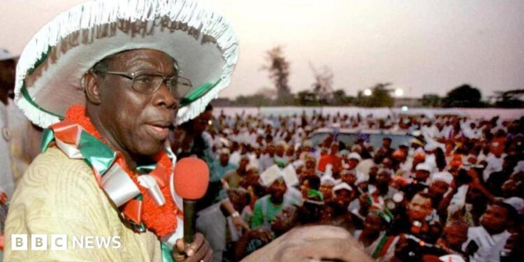 An archive picture of Olusegun Obasanjo from 1999. He is wearing a large white hat and addressing a crowd of people through a red microphone that he is holding.
