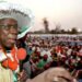 An archive picture of Olusegun Obasanjo from 1999. He is wearing a large white hat and addressing a crowd of people through a red microphone that he is holding.
