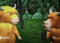A screengrab from the movie shows a yellow cow standing against poorly illustrated trees and talking to a brown bull that is holding what appears to be a black block