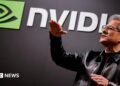 Nvidia CEO Jensen Huang standing in front of a screen with the company's name and logo, gesturing upward with his hands, wearing a black leather jacket over a black shirt.