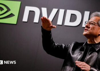 Nvidia CEO Jensen Huang standing in front of a screen with the company's name and logo, gesturing upward with his hands, wearing a black leather jacket over a black shirt.