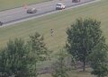 Highway camera view of a person running across a grassy field beside a multi-lane interstate with passing traffic.