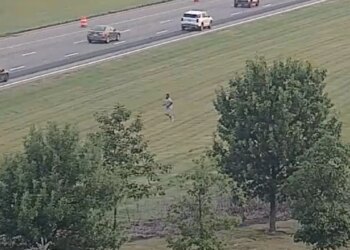 Highway camera view of a person running across a grassy field beside a multi-lane interstate with passing traffic.
