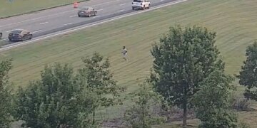 Highway camera view of a person running across a grassy field beside a multi-lane interstate with passing traffic.