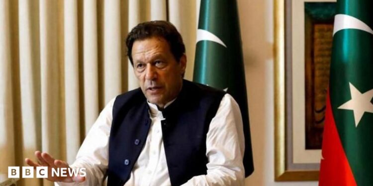 Former Pakistani Prime Minister Imran Khan speaks with Reuters during an interview, in Lahore, Pakistan March 17, 2023. He can be seen seated with his hand outstretched and speaking. Two Pakistani flags are behind him.