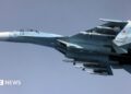 Image of a Russian Su-27, a whit and blue coloured fighter jet, airborne against a clear blue sky.