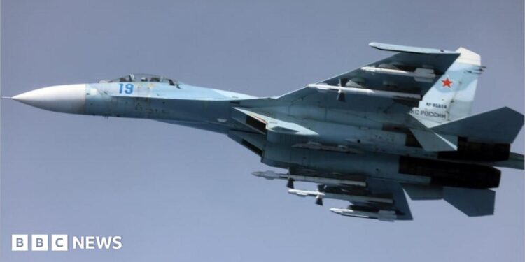 Image of a Russian Su-27, a whit and blue coloured fighter jet, airborne against a clear blue sky.