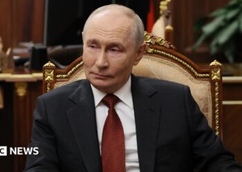 Russian President Vladimir Putin sits in a chair in an office, wearing a dark suit and red tie.