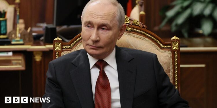 Russian President Vladimir Putin sits in a chair in an office, wearing a dark suit and red tie.
