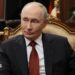 Russian President Vladimir Putin sits in a chair in an office, wearing a dark suit and red tie.
