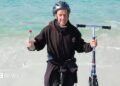 Bob Brown from Saltash, Cornwall stood on a beach in Australia having completed his challenge. He is stood just on the shoreline holding his scooter in one hand and a bottle of water in the other. He is wearing a black hoody and a black cycle helmet.
