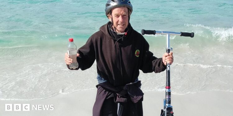 Bob Brown from Saltash, Cornwall stood on a beach in Australia having completed his challenge. He is stood just on the shoreline holding his scooter in one hand and a bottle of water in the other. He is wearing a black hoody and a black cycle helmet.