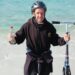 Bob Brown from Saltash, Cornwall stood on a beach in Australia having completed his challenge. He is stood just on the shoreline holding his scooter in one hand and a bottle of water in the other. He is wearing a black hoody and a black cycle helmet.
