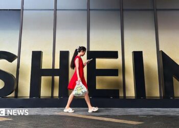 A woman in a red dress looks at her smartphone while walking past a Shein sign outside the company's office in Guangzhou, China.