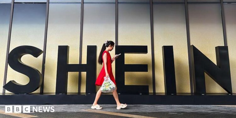 A woman in a red dress looks at her smartphone while walking past a Shein sign outside the company's office in Guangzhou, China.