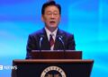 Lee Jae Myung speaks at a lectern during a Liberation Day ceremony. He wears a dark suit and glasses, in front of a blue backdrop.