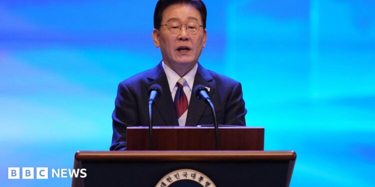 Lee Jae Myung speaks at a lectern during a Liberation Day ceremony. He wears a dark suit and glasses, in front of a blue backdrop.