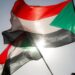 A Sudanese flag flutters in the wind against a backdrop of a bright blue sky - other Sudanese flags are in the background. The sun can be seen shining through the bottom of the flag.