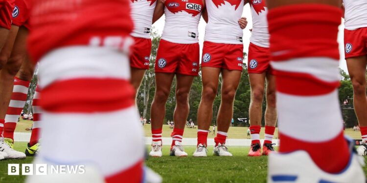 A pair of blurry legs in the foreground with a line of Sydney Swans players in the background