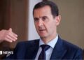 Bashar al-Assad pictured in 2016 speaking while gesturing with one hand. He is wearing a dark suit, white shirt and tie.