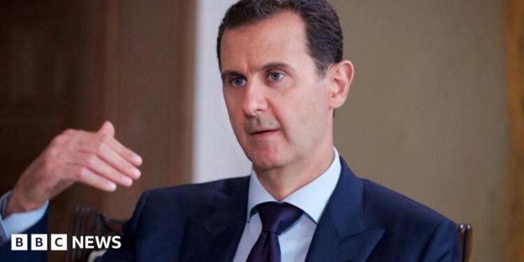 Bashar al-Assad pictured in 2016 speaking while gesturing with one hand. He is wearing a dark suit, white shirt and tie.