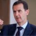 Bashar al-Assad pictured in 2016 speaking while gesturing with one hand. He is wearing a dark suit, white shirt and tie.