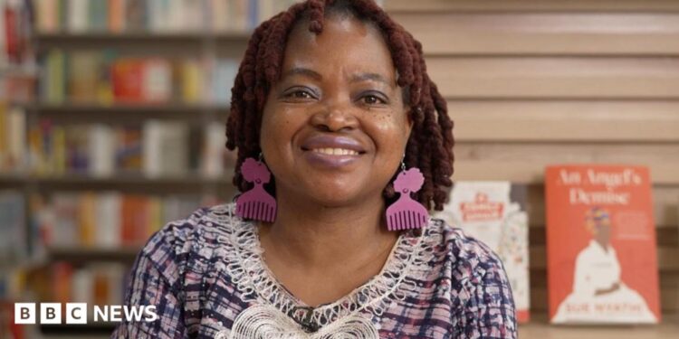 A woman with reddish-brown hair and pink comb-shaped earrings smiles at the camera, wearing a pink and blue checked top with white embroidered trim. She's in a bookshop, with blurred shelves of books on the left and a display featuring copies her books on the right.